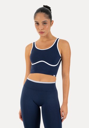 Kobieta z włosami upiętymi w kok, ubrana w granatowy sportowy krótki top i legginsy z wysokim stanem z białym obszyciem, stojąca na jednolitym tle.