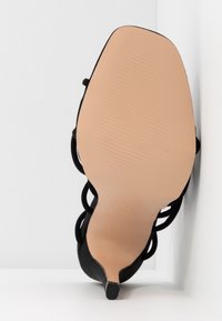 Sandale noire à talon haut avec des brides et une semelle beige, vue du dessous sur fond blanc.