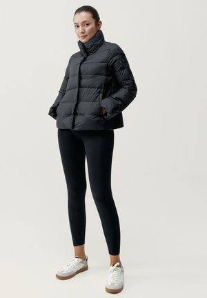 Zwarte pufferjas met een hoge kraag, voorzien van een doorgestikt design en zijzakken, gedragen over zwarte leggings en witte sneakers.