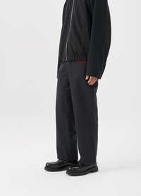 Svart zip-up jakke med elastisk kant, kombinert med løse svarte bukser. Skoene er skinnende svarte med en teksturert såle.