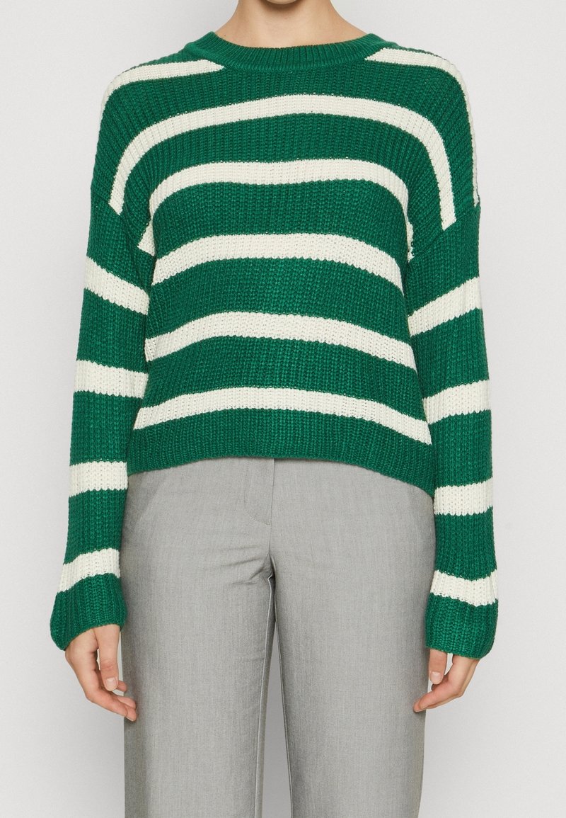 Maglione a righe verdi e bianche con una vestibilità rilassata, maniche larghe e collo rotondo, abbinato a pantaloni grigi chiari.