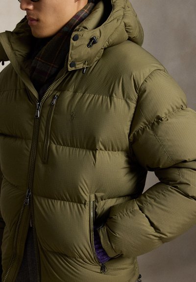 Personne portant une veste matelassée vert olive avec poches zippées et capuche, associée à une écharpe à carreaux sombre et des lunettes de soleil.