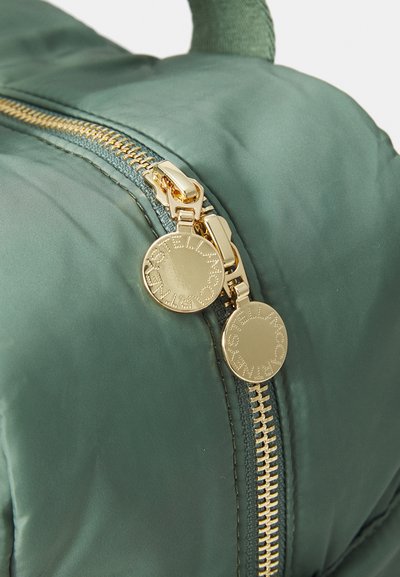 Rucsac verde din nylon cu un fermoar auriu și broșă rotundă, prezentând o textură netedă și cusături vizibile de-a lungul liniei fermoarului.
