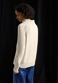 Cremfarbener Pullover mit einem strukturierten, übergroßen Kragen und geripptem Saum. Strickdesign mit offenen Nähten an den Schultern. Getragen mit Jeans.