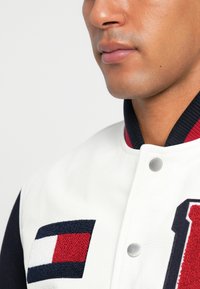 Giacca varsity in pelle bianca con maniche blu, patch del logo rosso e navy, bottoni in argento e colletto a coste. Tessuto testurizzato visibile sul patch.