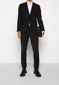 Czarny, dopasowany garnitur z klapami w szpic, zestawiony z jasnoróżową koszulą. Ubranie zawiera slim-fit spodnie i czarne skórzane buty oraz czarną torbę.