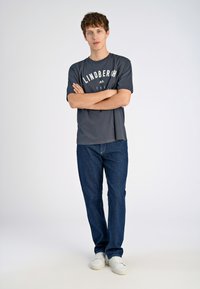 Harmaa lyhythihainen t-paita, jossa on valkoinen teksti "LINDBERGH" rinnassa, yhdistettynä sinisiin farkkuihin ja valkoisiin lenkkareihin. Yksivärinen tausta.