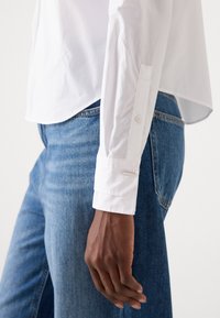 Witte langslijp shirt met een puntig kraag, enkele knoopmanchetten en een subtiele logo op de borst, gecombineerd met blauwe denim jeans.