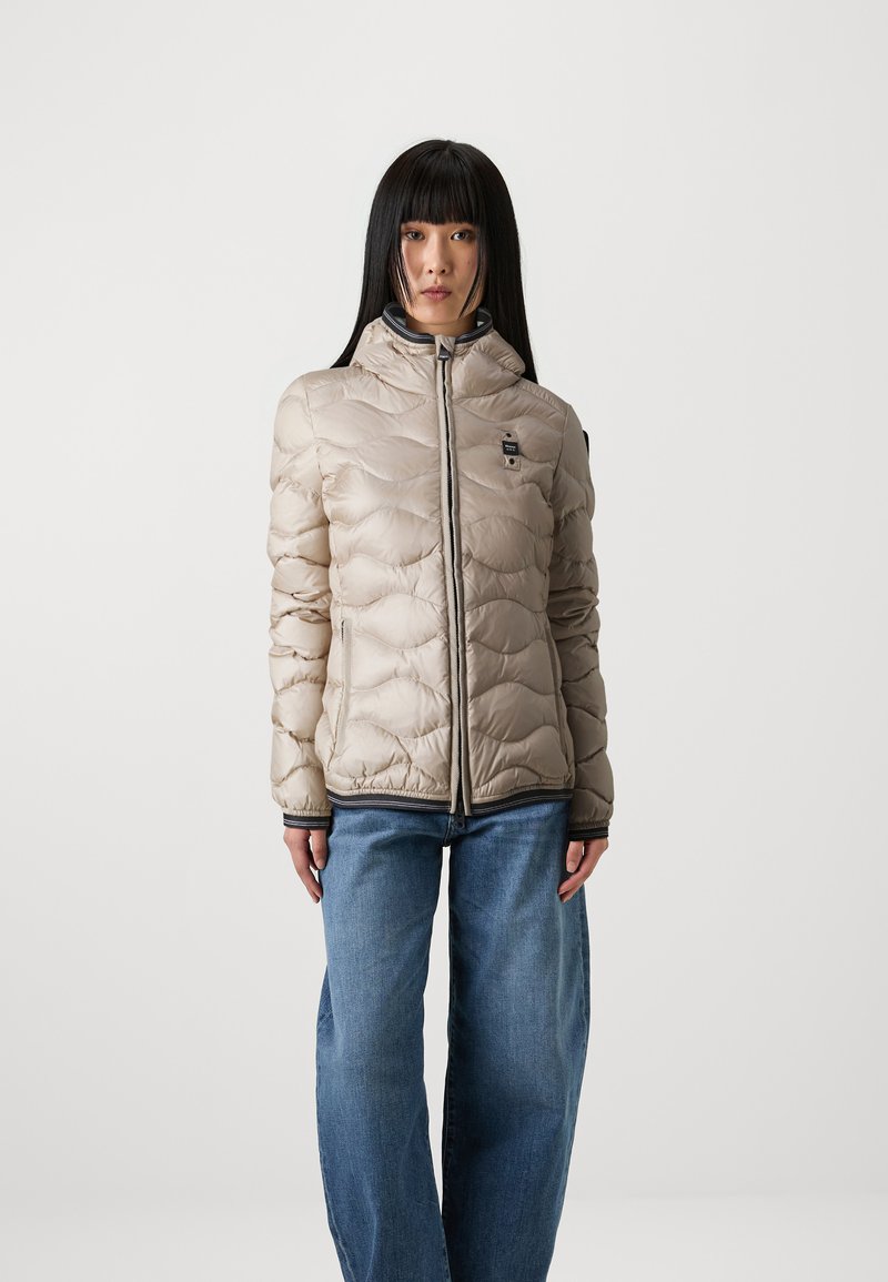 Giacca a vento beige con design trapuntato, colletto alto e chiusura con zip. Presenta una rifinitura grigia e una toppa con logo sul lato sinistro del petto.