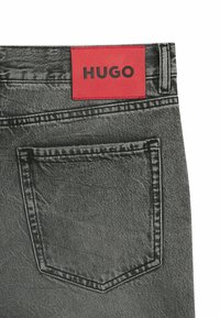 Jeans in denim grigio con etichetta in pelle rossa con scritta "HUGO" in testo nero, strutturati con una tasca posteriore e cuciture a contrasto.