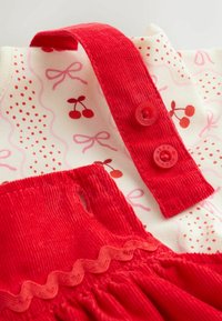 Red corduroy straps with buttons attached to a white fabric patterned with red cherries and pink bows and dots.