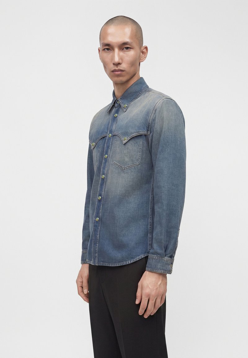 Mand iført en falmet blå denimskjorte med brystlommer og sorte bukser, stående med fronten mod kameraet med neutralt udtryk.