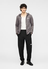 Γκρι μπουφάν με φερμουάρ και κουκούλα· λευκό τοπ από μέσα. Μαύροι cargo παντελόνια με τσέπες· μαύρα αθλητικά παπούτσια. Λογότυπο EA7 στο μπουφάν και τα παντελόνια.