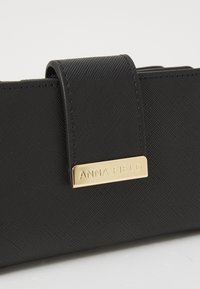 Portefeuille noir texturé en matériau synthétique, avec une plaque de marque dorée et une fermeture à rabat. Design simple et rectangulaire.