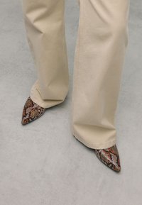 Spitz zulaufende Schuhe im strukturierten, mehrfarbigen Schlangenprint-Muster. Getragen mit beigen, weiten Hosen, die kunstvolle Details und lebendige Akzente zeigen.
