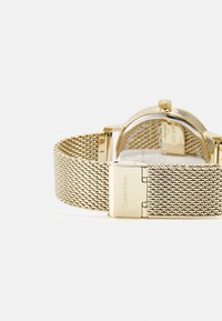 Die goldene Mesh-Armbanduhr verfügt über ein rundes Gehäuse, eine mit "Calvin Klein" gravierte Schließe und einen transparenten Rücken, der das Uhrwerk zeigt.