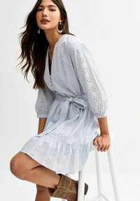 Lichtblauwe jurk met oogjesbloemmotief, V-haak, poffende mouwen, ceintuur op de taille en gerafeld zoom. Model gezeten op een witte kruk.