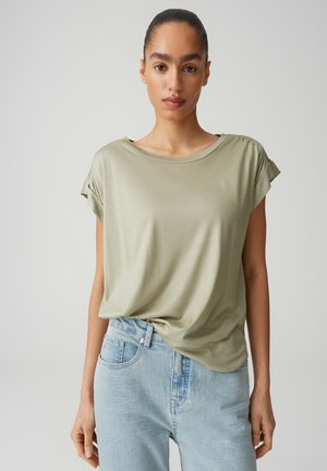 Jeune femme aux cheveux tirés en arrière portant un haut ample à manches courtes de couleur vert olive clair et un jean bleu clair, debout devant un fond uni.