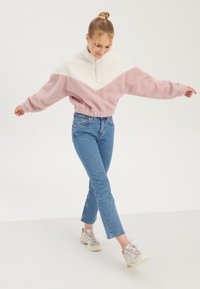 Pullover chaud zippé, en bloc de couleurs blanc et rose clair, avec une finition douce et texturée. Associé à un jean droit bleu et des baskets.