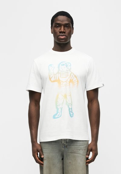 Άνδρας που φορά λευκό μπλουζάκι με πολύχρωμο γραφικό αστροναύτη που κυματίζει, στέκεται μπροστά σε απλό λευκό φόντο.