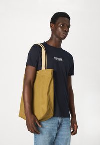 Borsa tote in tela di colore giallo senape, con lunghe maniche crema e una forma strutturata. Indossata da un modello con una t-shirt blu navy e jeans chiari.