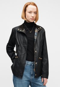 Γυναίκα με κοντά κόκκινα μαλλιά, φορούσα μαύρο πουλόβερ με ζιβάγκο, μαύρο μπουφάν με φερμουάρ και χρυσά κουμπιά, και μπλε τζιν, στέκεται μπροστά σε απλό φόντο.