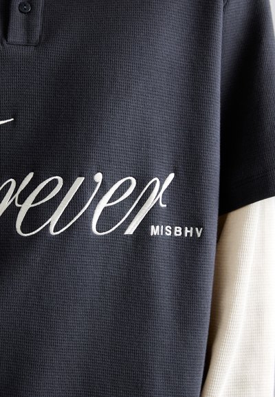 Polo de color azul marino texturizado con "forever" y "MISBHV" bordados en blanco en el lado izquierdo; presenta una tapeta de dos botones.