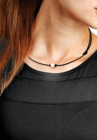 Collier en cordon noir avec une seule perle crème, con�çu avec des nœuds ajustables pour personnaliser la longueur. Porté sur un haut noir.