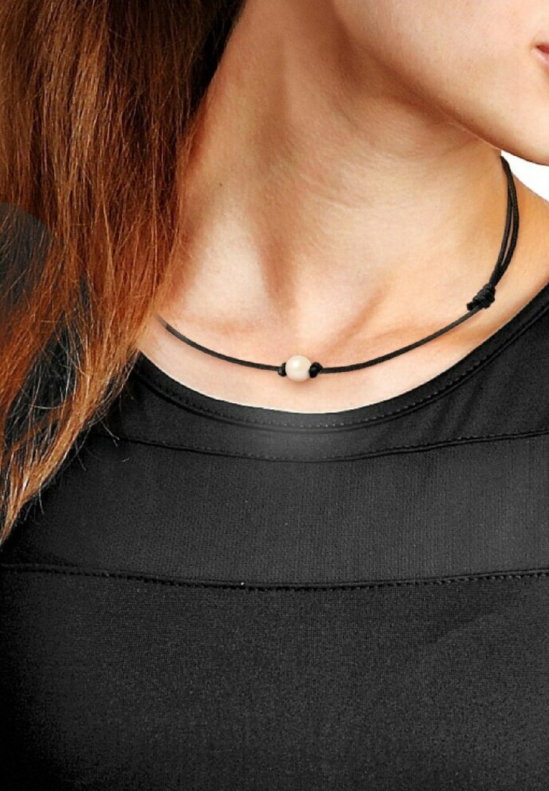 Collier en cordon noir avec une seule perle crème, conçu avec des nœuds ajustables pour personnaliser la longueur. Porté sur un haut noir.