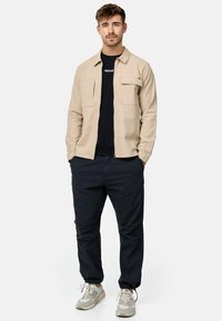 Chemise beige à boutonnage avec deux poches poitrine, portée sur un t-shirt noir, associée à un pantalon marine et des baskets gris clair.