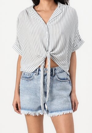 Donna che indossa una camicia a maniche corte bianca e grigia a righe verticali annodata sul davanti e pantaloncini di jeans azzurri chiari sfrangiati con motivi in strass.