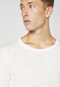 Balta tekstūruota marškinėliai su apvaliu iškirpimu. Šiek tiek permatomas audinys, minkšta išvaizda. Modelis nusisukęs į šoną, atidengdama kaklą ir petį.