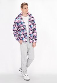 Jachetă colorată cu un model de flamingo în roz, albastru și negru. Pantaloni jogger gri și adidași albi. Modelul stă relaxat, cu mâinile în buzunare.