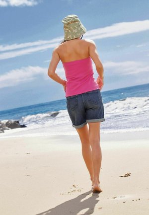 Femme en top tube rose, short en denim et chapeau bob vert marchant pieds nus sur une plage de sable près des vagues de l'océan sous un ciel bleu.