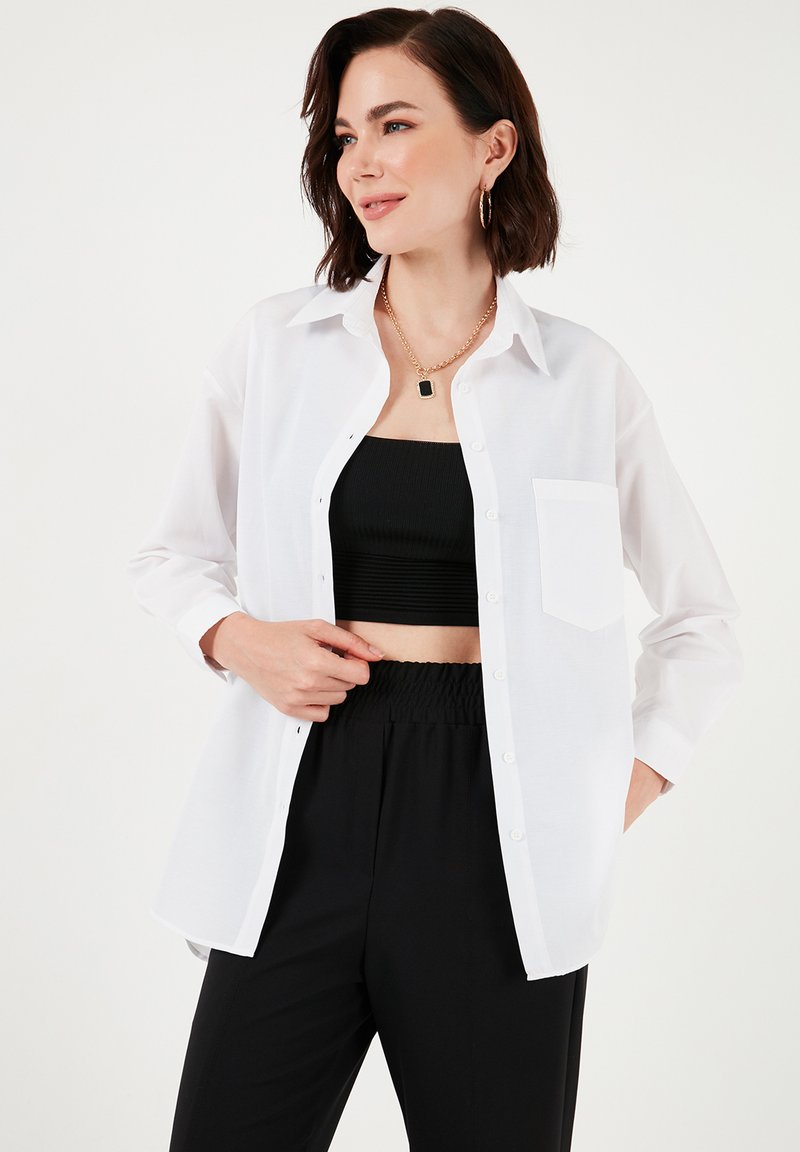 Camicia bianca con maniche lunghe, dotata di una singola tasca sul petto, abbinata a un top corto nero e pantaloni con cintura elastica.