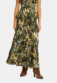 Abito maxi con vita a peplum, vivace motivo astratto verde e oro, e silhouette fluida, abbinato a stivali con tacco marroni.