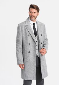 Abrigo de lana gris en patrón de espiga con diseño de doble botonadura, seis botones negros, solapas muesca y bolsillos laterales. Llevado sobre un chaleco gris y una camisa blanca.