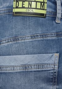 Modrá denimová látka má viditelný vzor šití. Obdélníková etiketa v jasně zelené a černé barvě nese nápis "Cecil Denim." Klasický design kapes.