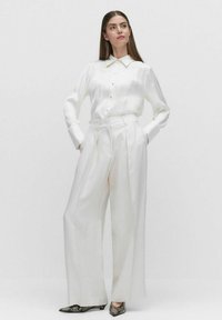 Witte zijden blouse met een puntkraag en knoopsluiting, gecombineerd met hoge, wijde witte broeken en gepatineerde hakken.