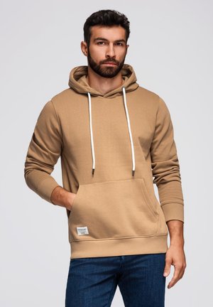 Beżowa bluza z kapturem z przednią kieszenią, białymi sznurkami i ściągaczami, zestawiona z ciemnoniebieskimi dżinsami. Gładka faktura materiału.