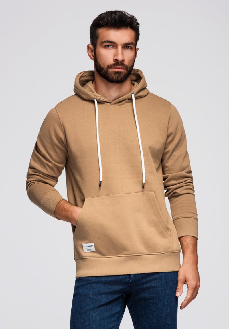 Hnědá mikina s kapucí, přední kapsou, bílými tkaničkami a pružnými manžetami, v kombinaci s tmavě modrými džíny. Hladká textilie.
