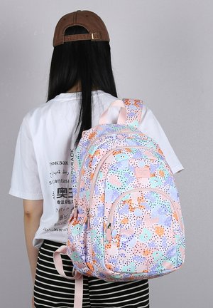 Jonge persoon met lang zwart haar, bruine pet, wit grafisch T-shirt, gestreepte broek, dragend een rugzak met pastelkleurig multicolor patroon.