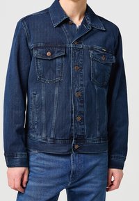 Homme portant une veste en denim bleu foncé avec des poches poitrine boutonnées et un jean bleu assorti, sur un fond uni.