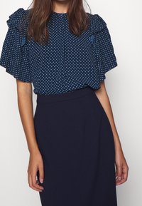 Marinblå blus med polka prickar och volangärmar, kombinerad med en enfärgad mörk kjol. Slät tygtextur, avslappnad design.