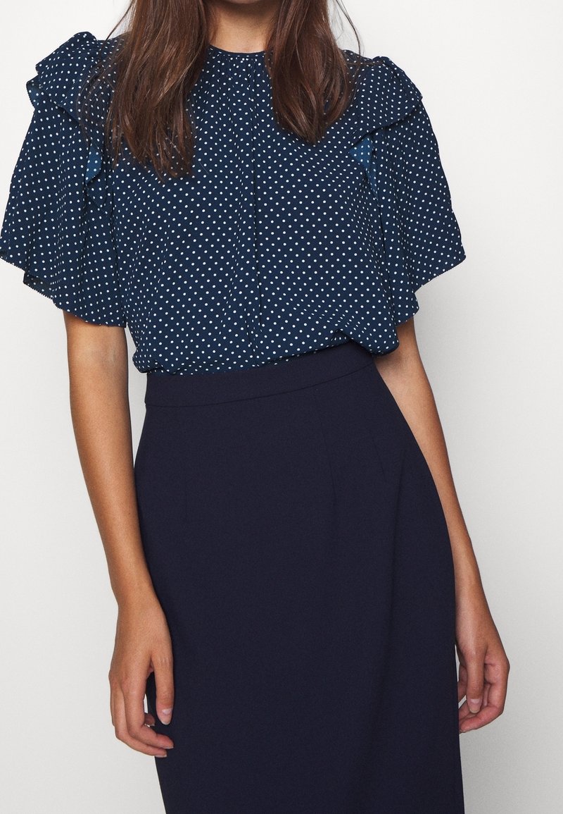 Marinblå blus med polka prickar och volangärmar, kombinerad med en enfärgad mörk kjol. Slät tygtextur, avslappnad design.