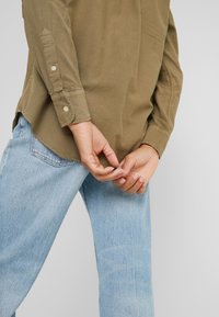 Olivově zelená košile na knoflíky s ohrnutými rukávy, texturovaná tkanina, spárovaná s světle modrými džíny v tapered střihu a s jemným praním.