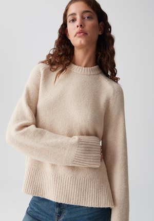 Vrouw met golvend bruin haar, gekleed in een beige wollen trui en blauwe jeans, staande tegenover een effen lichtgrijze achtergrond.