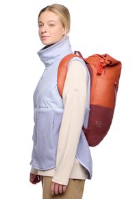 Chaleco aislante azul claro con una sección inferior texturizada morada, llevado sobre una camiseta de manga larga color crema; una mochila naranja con parte superior enrollable al hombro.