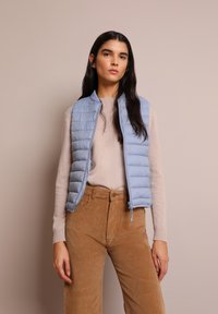 Gilet imbottito azzurro chiaro con design a trapunte verticali, chiusura con zip e un maglione in maglia beige a contrasto. Pantaloni in corduroy nei toni della terra.