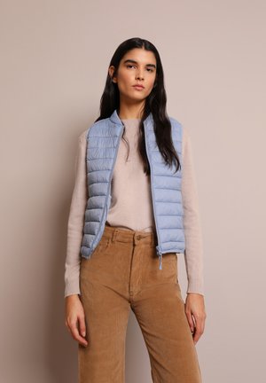 Gilet imbottito azzurro chiaro con design a trapunte verticali, chiusura con zip e un maglione in maglia beige a contrasto. Pantaloni in corduroy nei toni della terra.